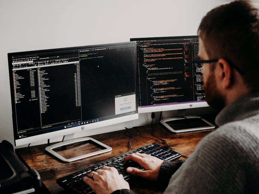 A person with glasses and a beard is seated at a wooden desk, working on dual computer monitors displaying lines of programming code in a dark-themed editor. The left monitor shows a file directory an