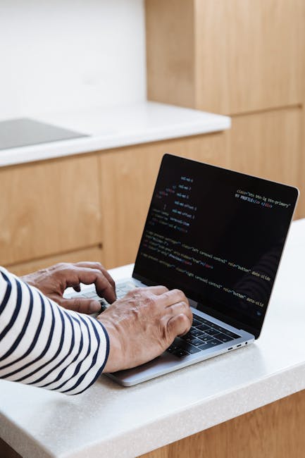 A person with glasses and a beard is seated at a wooden desk, working on dual computer monitors displaying lines of programming code in a dark-themed editor. The left monitor shows a file directory an
